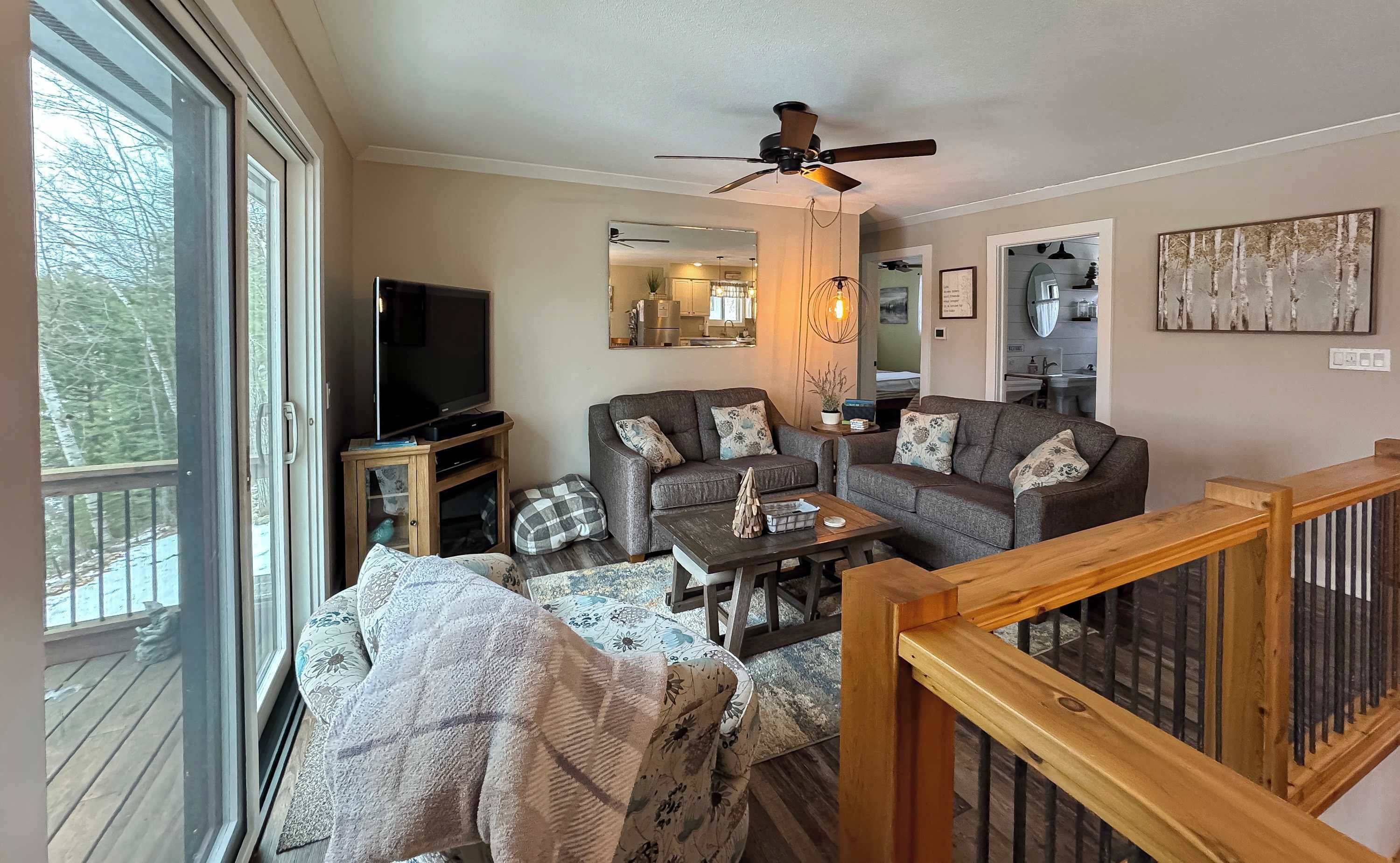 A living room with a sofa, a television, a ceiling fan, and a view of the outdoors.