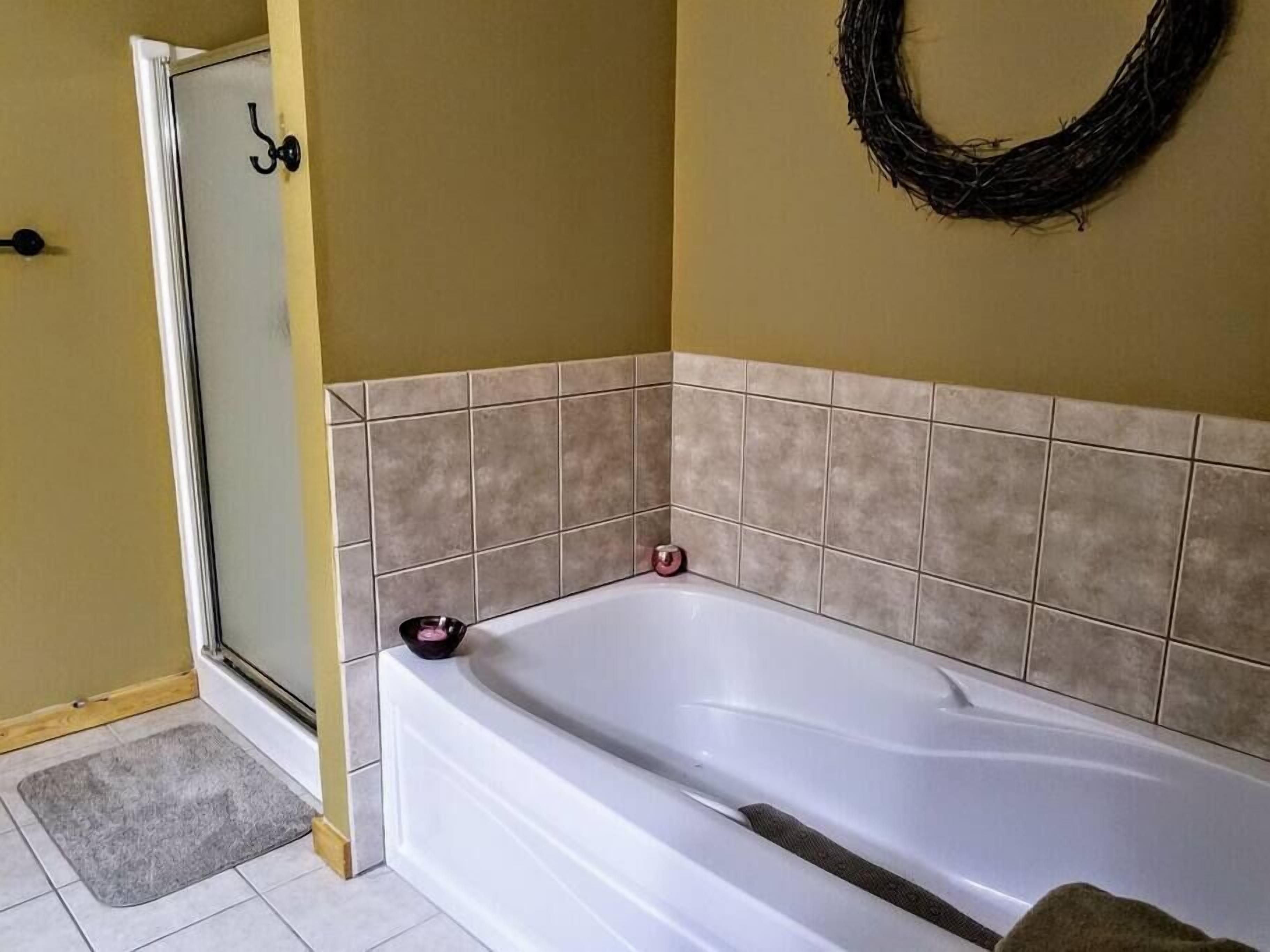 A bathroom with a white bathtub, beige tiled walls, a shower area, and a gray mat on the floor.