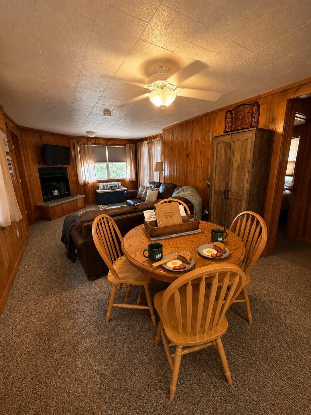 A cozy cabin interior with a dining table set for breakfast, a fireplace, and a television mounted on the wall.