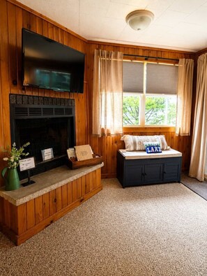 Een kamer met een open haard, een televisie erboven, een raam met gordijnen, een bank met kussens en een kleine tafel met een bord.'