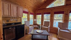 A cozy cabin interior with wooden walls, a kitchen area, and seating by large windows. - Hilltop - Bouchard Shores Resort (Minocqua)