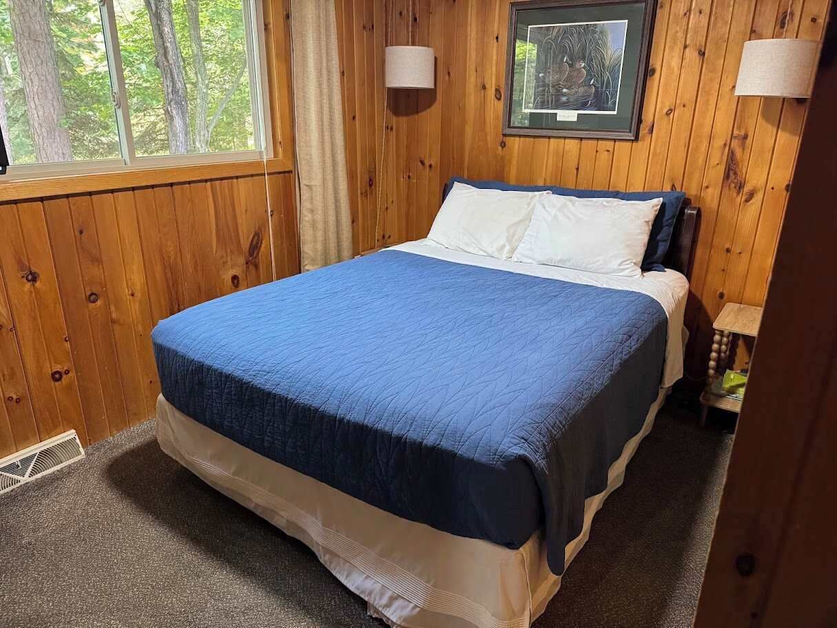 A wooden-paneled room with a bed, a window, and a framed picture on the wall.