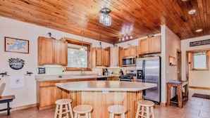 A modern kitchen with wooden cabinets, a central island with stools, stainless steel appliances, and a large window. - Eikedahls' Shoreview Lake House (Minocqua)