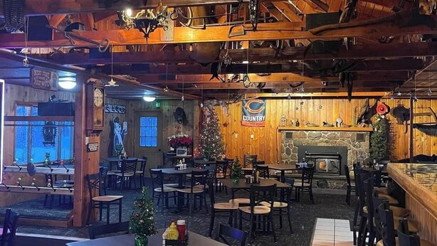 Een rustiek interieur met houten balken, een open haard en een kerstboom. Tafels en stoelen staan klaar voor een diner.