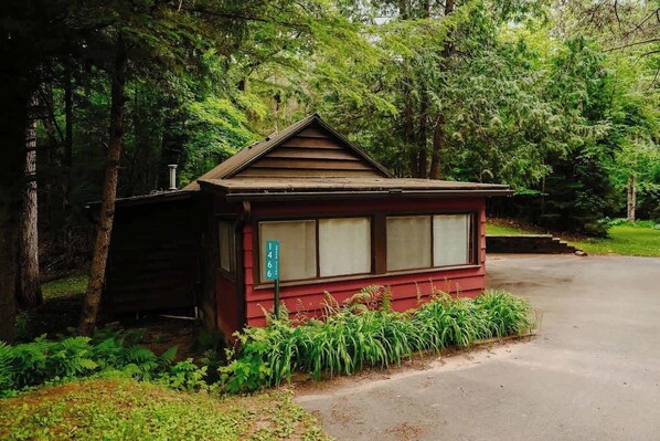 A small, rustic cabin with a sloped roof, surrounded by dense greenery and trees. - Old Stoney's Perch - Big Arbor Vitae Lake (Woodruff)