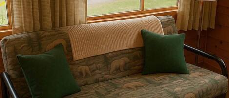 A cozy room with a sofa featuring a bear pattern, two green pillows, and a view of a park through a window.