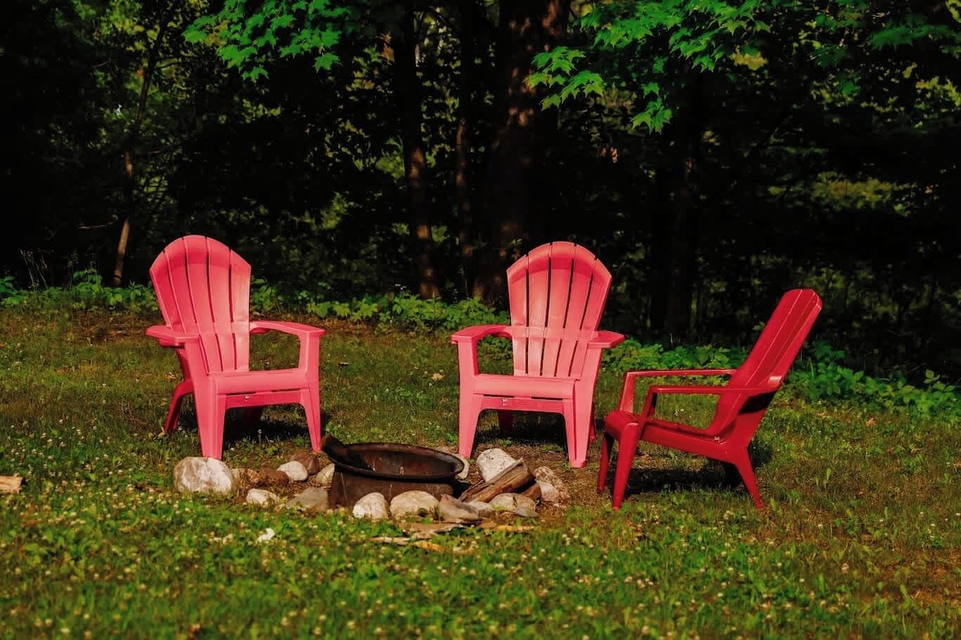 Two red Adirondack chairs and a fire pit in a grassy outdoor area with trees in the background.