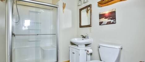 Un bagno con box doccia in vetro, un lavandino bianco, un water e una piccola mensola in legno con uno specchio e un quadro.