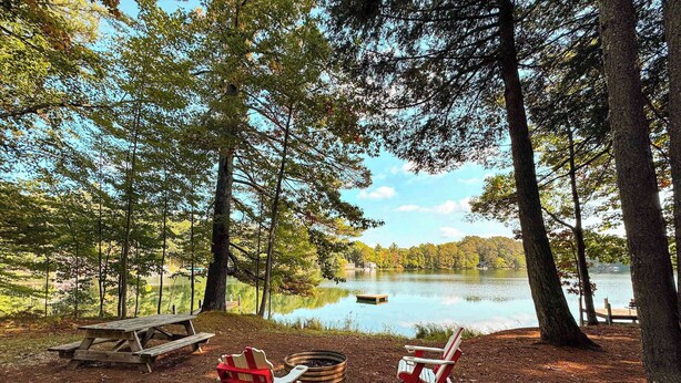 Ein Campingplatz am Seeufer mit zwei roten Adirondack-Stühlen, einem Picknicktisch und einem kleinen Steg.