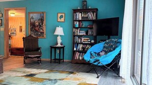 Een woonkamer met een wand in groenblauw, een boekenkast, een televisie, een blauwe stoel en een vloerkleed met een patroon.
