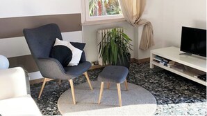 Ein modernes Wohnzimmer mit einem grauen Sessel, einem kleinen Hocker, einer weißen TV-Konsole und einer großen Topfpflanze.
