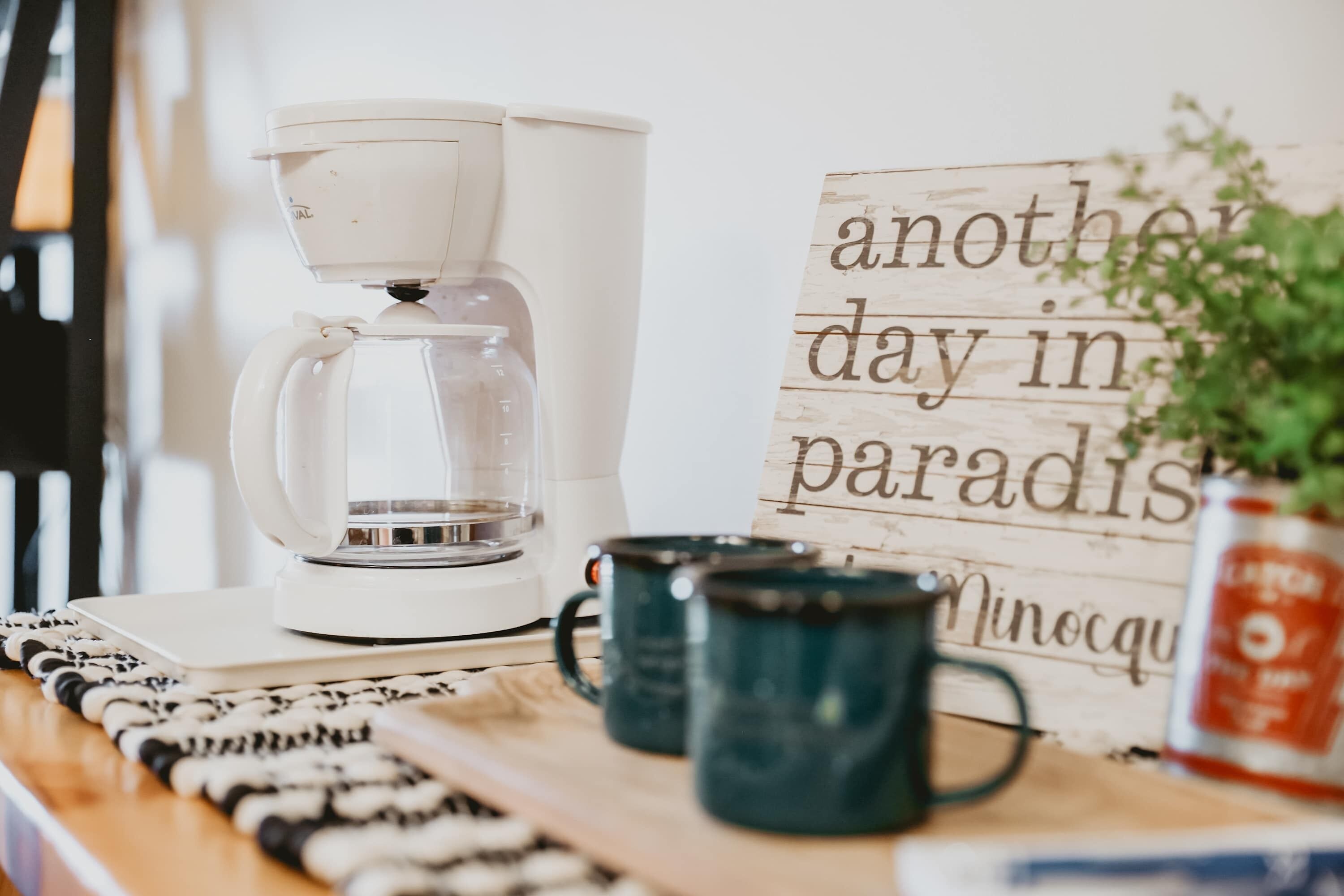 A coffee maker on a table with a sign reading "another day in paradise" and two mugs.