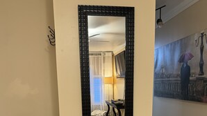 A mirror with a black frame reflecting a room with a bed, a lamp, and a window with curtains.
