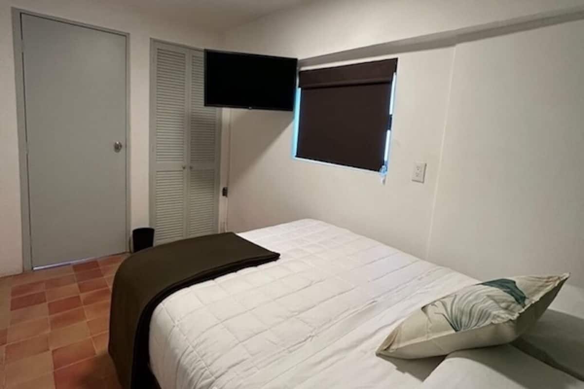 A bedroom with a bed, a flat-screen TV mounted on the wall, a window with blinds, and a door.