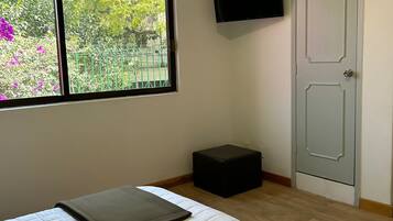 Quarto com cama, janela com vista para vegetação, televisão presa na parede e porta.