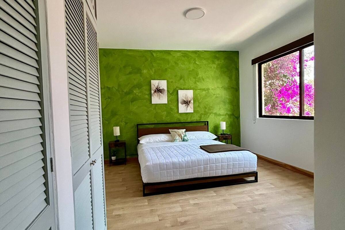 A bedroom with a bed, green wall, window with pink flowers, and wooden flooring.