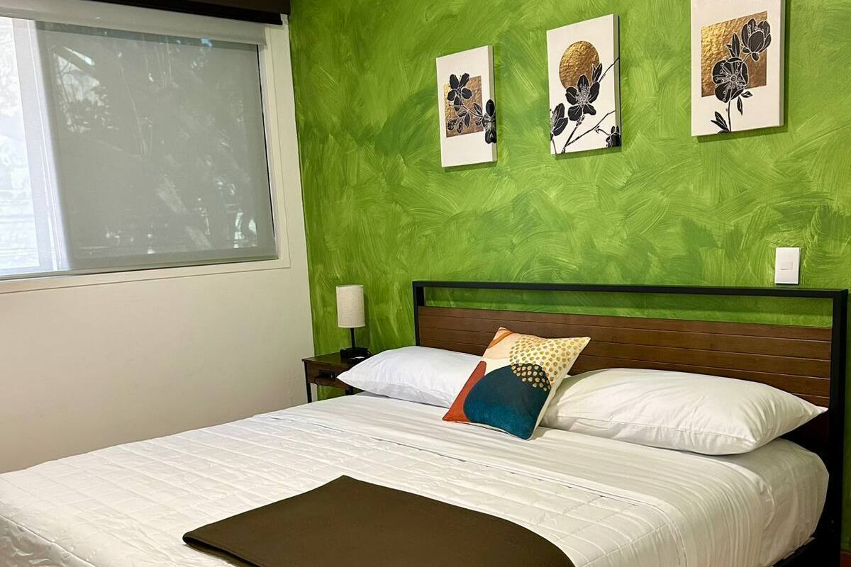 A bed with a wooden frame, white bedding, and a green wall with three framed floral artworks.