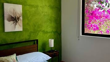 Un dormitorio con pared verde, cama con ropa blanca, mesita de noche con lámpara y ventana con flores rosadas.