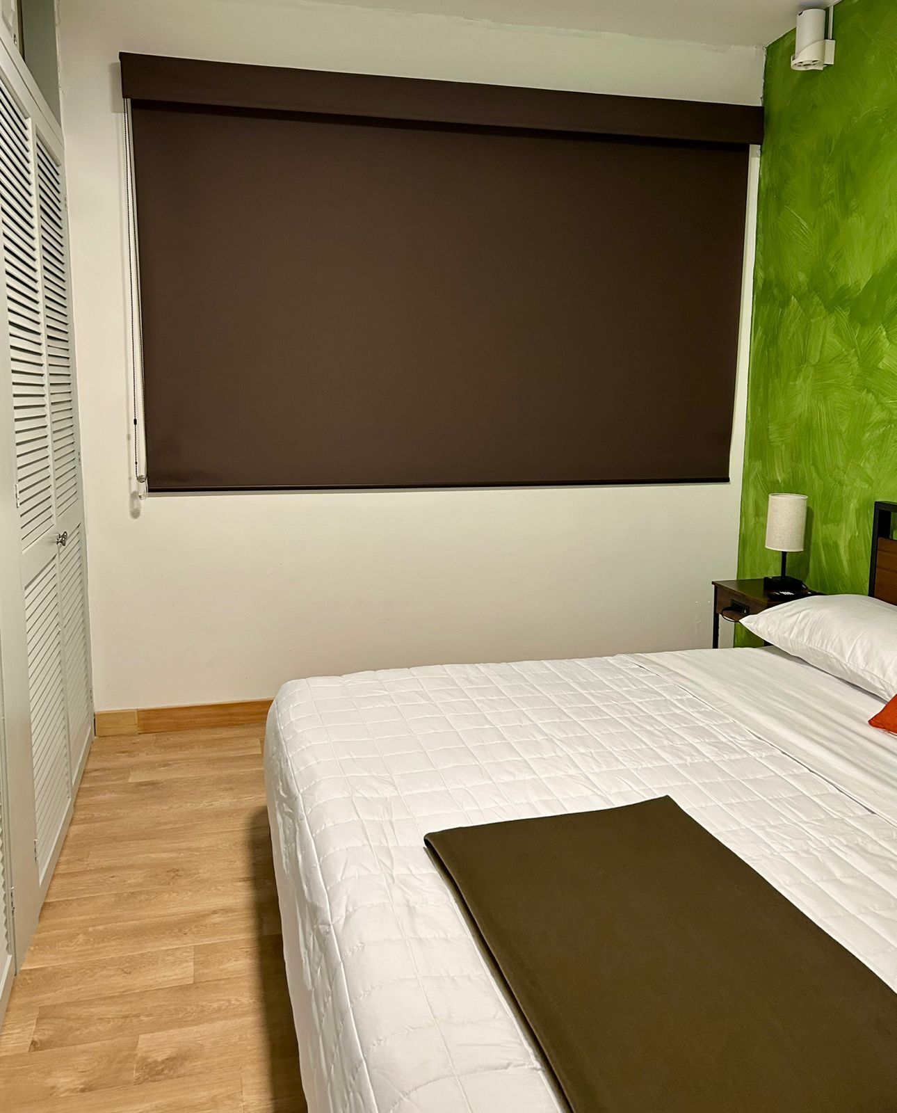 Une chambre d’hôtel avec un lit, une grande fenêtre avec un store, un mur d’accent vert, une table de chevet avec une lampe et un plancher en bois.