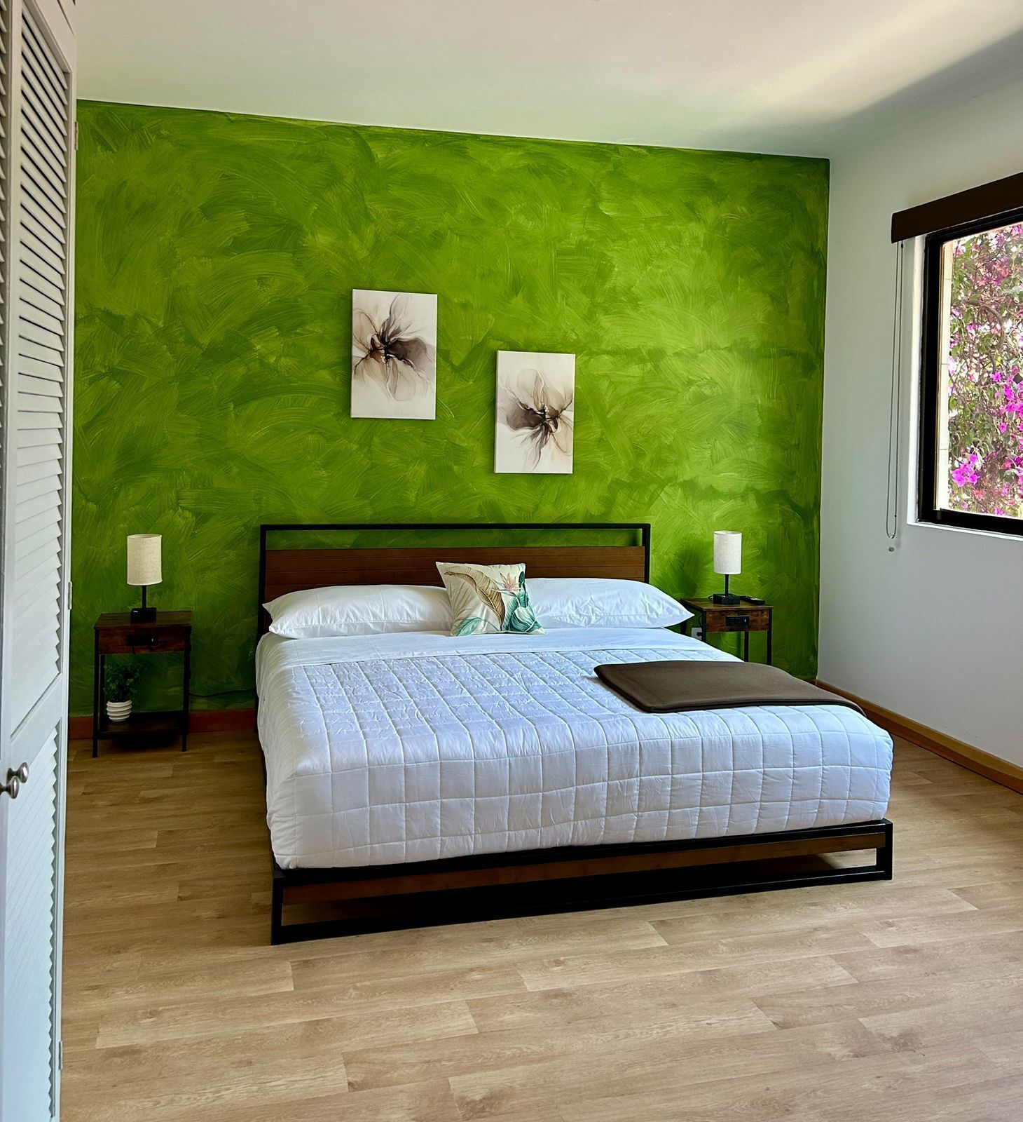 A bedroom with a green wall, a wooden bed frame, white bedding, two wall art pieces, and a window with a view of pink flowers.