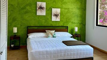A bedroom with a green wall, a wooden bed frame, white bedding, two wall art pieces, and a window with a view of pink flowers.