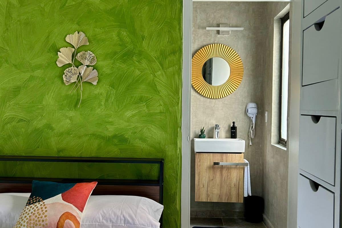 A bedroom with a bed, a green wall, a mirror, and a bathroom visible through an open door.