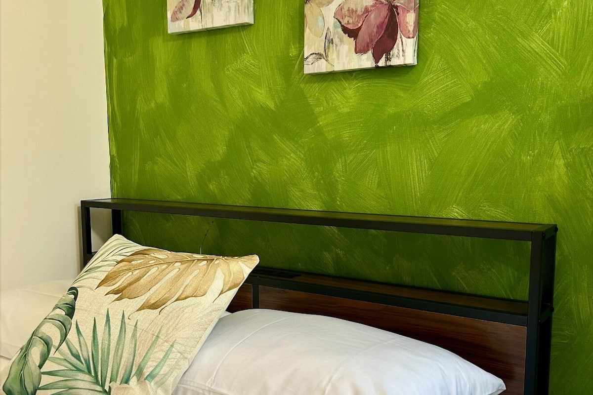 A bed with a white pillow and a floral-patterned pillow, a green wall, and two paintings of flowers.