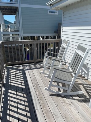 A wooden deck with white rocking chairs, a white railing, and a blue building in the background. - The Compass OKI: Beautiful Beach House on OKI, NC- 250 feet from access (Oak Island)