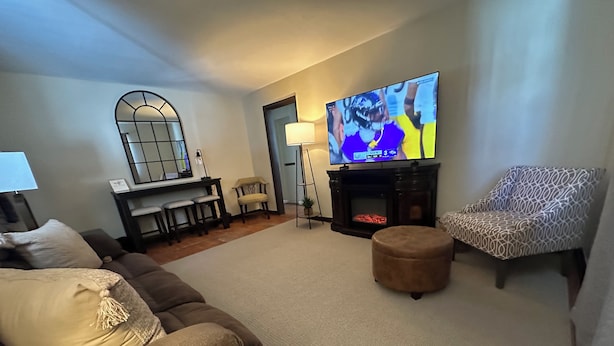 A living room with a sofa, ottoman, patterned armchair, TV, fireplace, and a dining area with chairs and a table. - Super Clean 2BR House: Quiet, Safe, Near Hwy, Big TV, Nightlife, Porch, by River (Beaver)