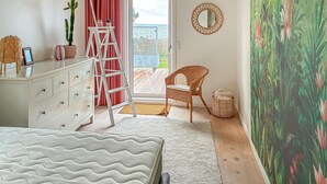 Ein Schlafzimmer mit Bett, Kommode, einer Himmelsleiter, Stuhl, Korb und einem großen Fenster mit Blick ins Freie.