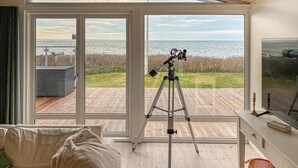 Ein Zimmer mit einem großen Fenster mit Blick auf eine Terrasse und Wasser. Auf der Terrasse steht ein Teleskop.
