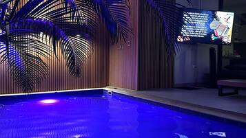 A swimming pool with blue lighting, surrounded by palm trees and a television screen displaying a program.