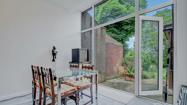 Een moderne eetruimte met een glazen tafel en houten stoelen, een televisie op een standaard, en uitzicht op een bakstenen gebouw en groen buiten.