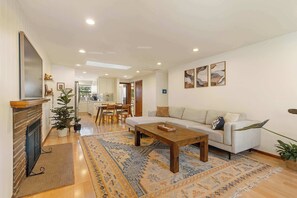 A spacious living area with a sofa, coffee table, and a fireplace. There is a dining area with chairs and a kitchen in the background. - 3BR Seattle Home w/ Spacious Yard | Near Northgate (Seattle)