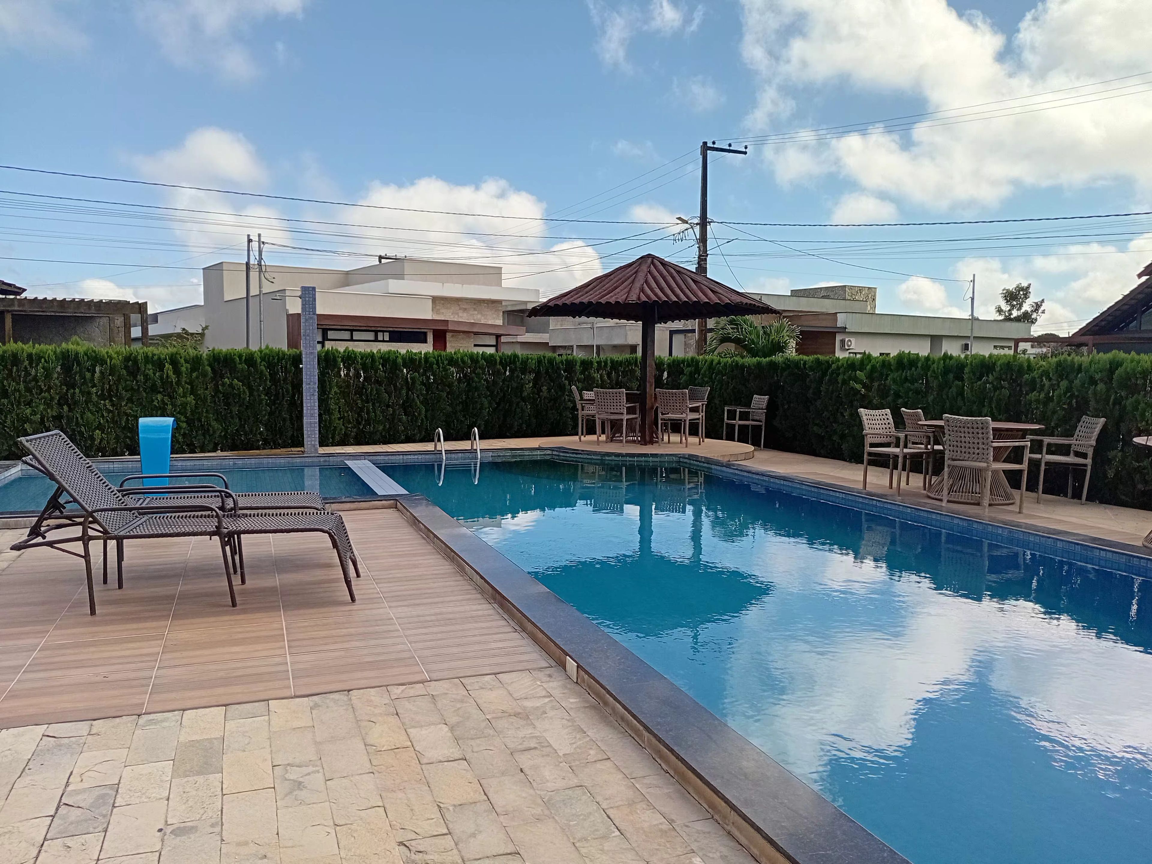 Ein Swimmingpool mit Liegestühlen und einer überdachten Sitzgelegenheit, umgeben von Hecken und Gebäuden.