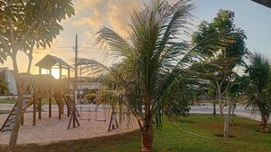 A playground with swings and a slide, surrounded by palm trees and a grassy area.