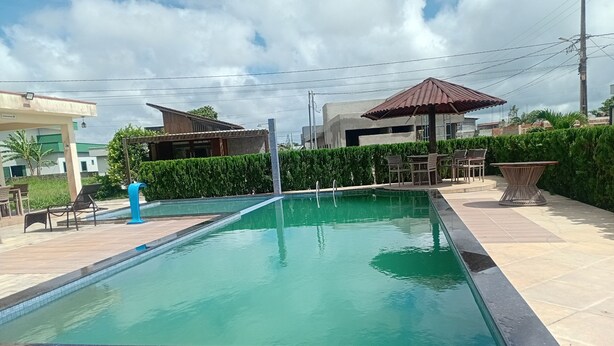 A rectangular swimming pool with a tiled deck, surrounded by greenery and a few outdoor seating areas.