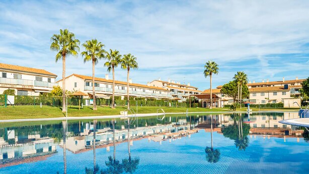 A swimming pool with clear blue water, surrounded by palm trees and modern buildings with a Mediterranean architectural style.