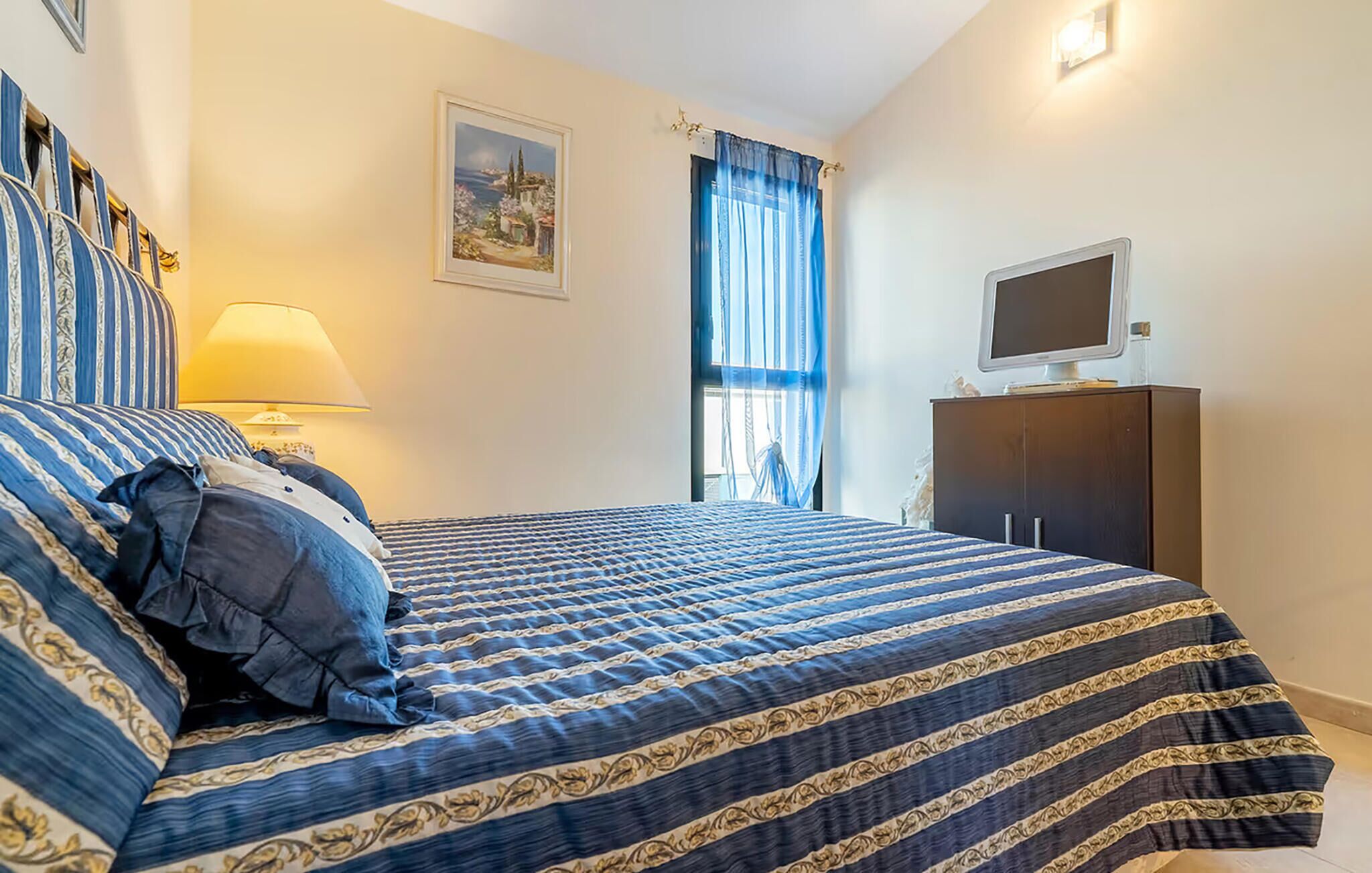 Une chambre à coucher avec un lit, une table de chevet, une lampe, un téléviseur posé sur un meuble et une fenêtre avec des rideaux.