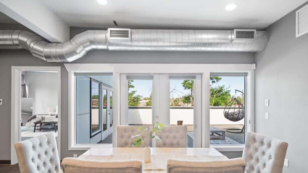 A modern dining area with a large table and chairs, large windows, and an air conditioning duct above.