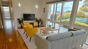 Un salon moderne avec un canapé gris, des fauteuils jaunes, un téléviseur à écran plat et une grande fenêtre donnant sur l’extérieur.