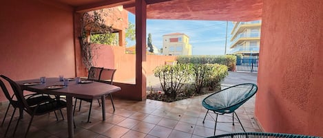 A covered patio with a table and chairs, a small garden, and a view of buildings in the background.