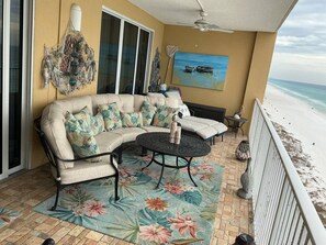 A balcony with a sofa and a round table, a decorative hanging, and a painting of a beach scene. - Tropic Winds 905 (Panama City Beach)