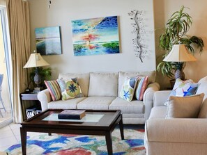 A living room with a sofa, a coffee table, and two lamps. There are two paintings on the wall and a plant on the right. - Tropic Winds 905 (Panama City Beach)