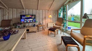 Une pièce chaleureuse avec un plancher en bois, un téléviseur fixé au mur, une table avec des chaises et une fenêtre donnant sur un paysage verdoyant.