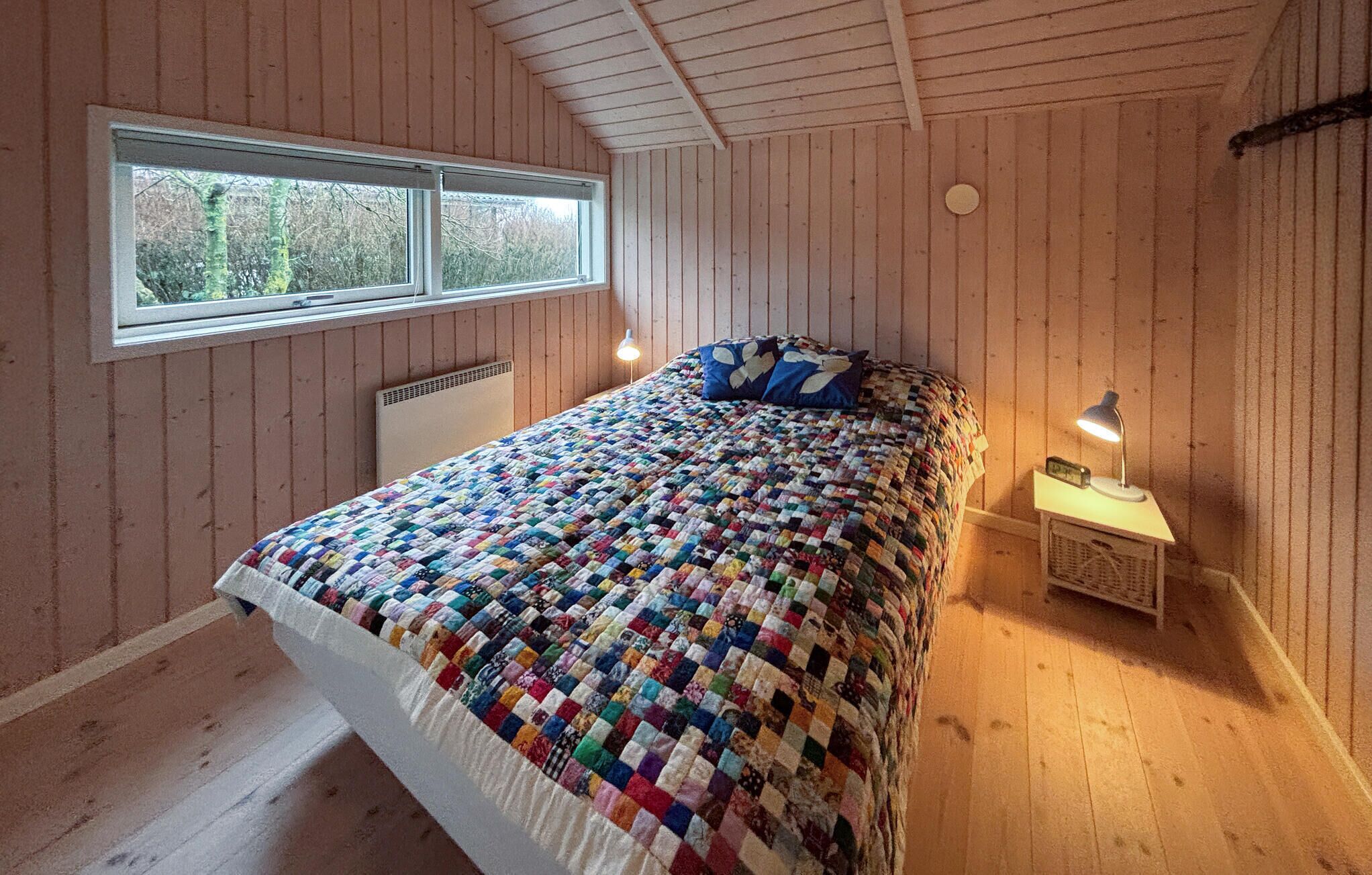 A bedroom with a quilted bedspread, a window, a bedside lamp, and a radiator.