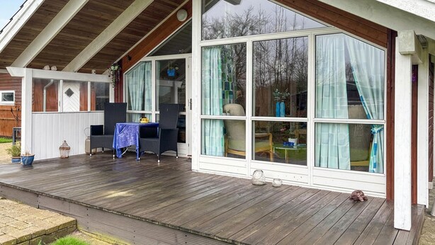 Une véranda couverte avec une terrasse en bois, une table et des chaises, et de grandes fenêtres avec des rideaux.