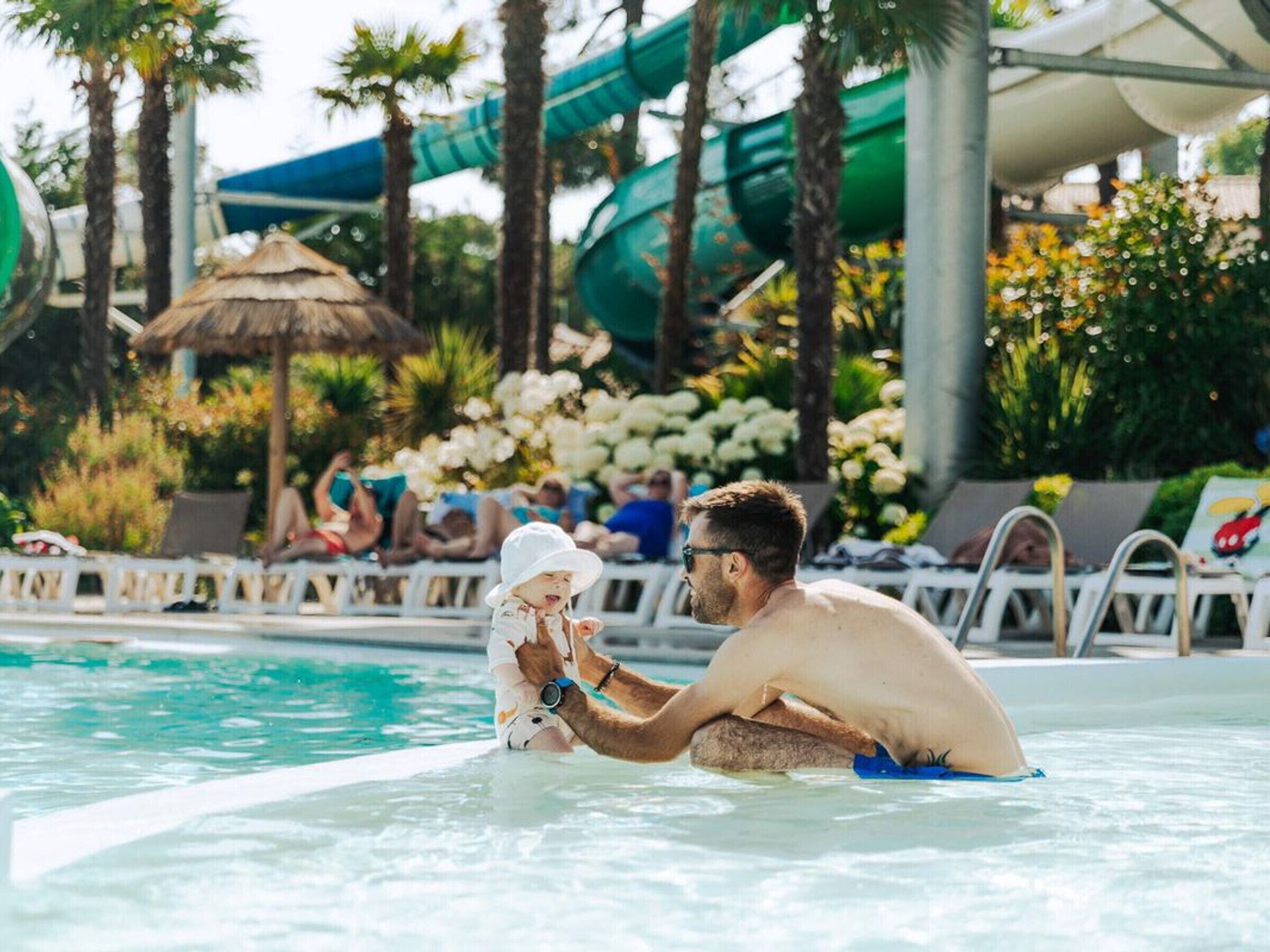 A man in a swimming pool with a child, surrounded by palm trees and a water slide.