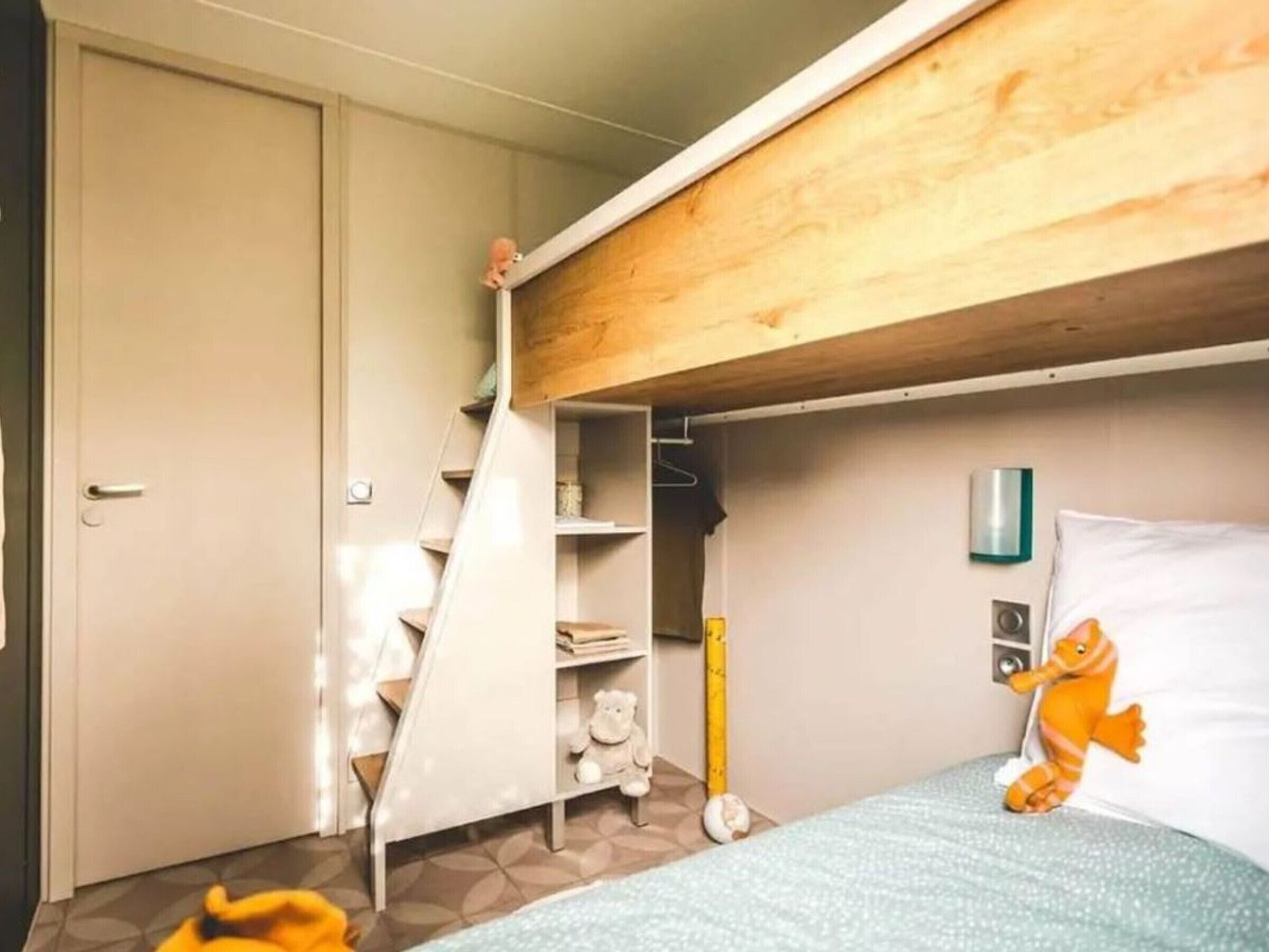 A bedroom with a bunk bed, a wardrobe, a shelf with toys, and a bed with a pillow and stuffed animal.