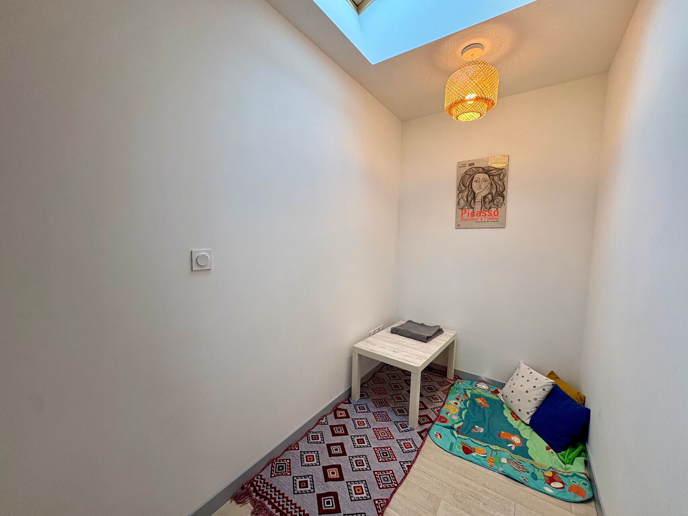 Un couloir étroit avec une lucarne, une petite table sur laquelle est posé un vêtement plié, un tapis coloré et un lit avec des oreillers.